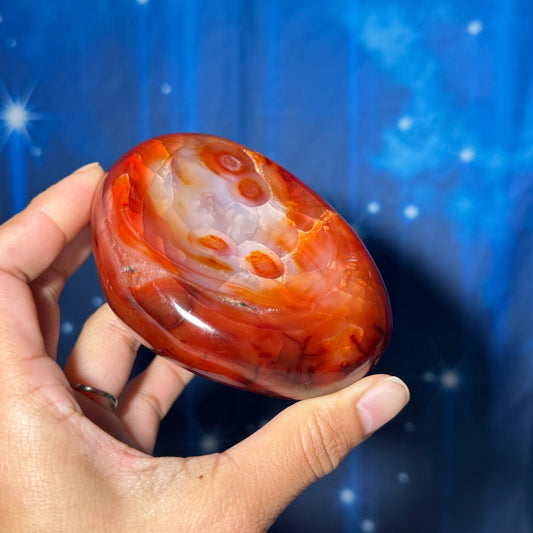 May include: A red carnelian crystal with white and orange banding. The crystal is smooth and polished and has a rounded shape.
