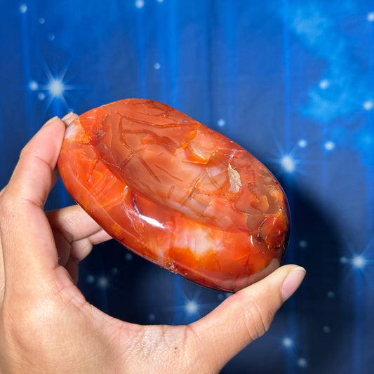 May include: A smooth, polished carnelian stone with a deep orange and red color. The stone is shaped like a shallow bowl and has a natural, organic pattern.