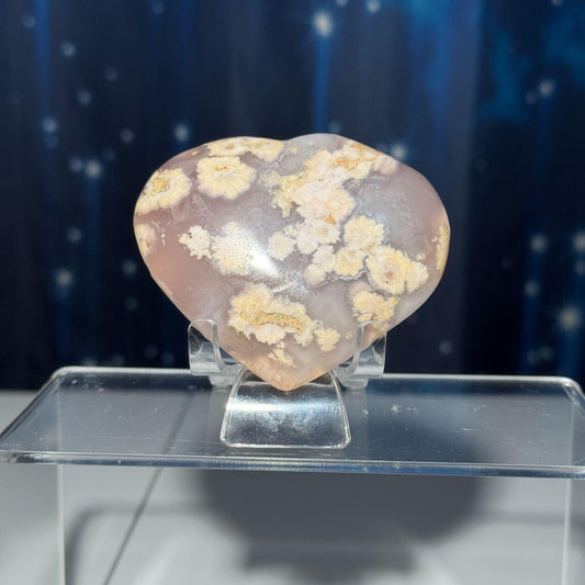 May include: A heart shaped pink and white agate crystal with a white, cloudy pattern. The crystal is on a clear plastic stand.