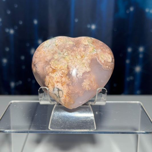 May include: A heart shaped pink agate crystal with white and green inclusions. The crystal is sitting on a clear acrylic stand.